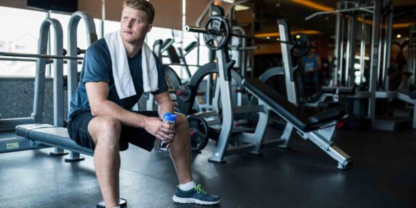 A man seated on a bench inside a gym, surrounded by exercise equipment and a focused atmosphere. - Fitsse