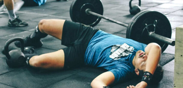 A man resting on the floor of a CrossFit gym, surrounded by workout equipment and a focused training environment. - Fitsse