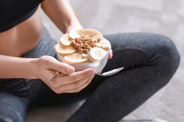 A woman holds a bowl of yogurt topped with sliced bananas and nuts, showcasing a healthy snack option. - Fitsse