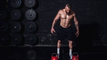 A man stands confidently, holding a kettlebell in each hand, ready to engage in a powerful workout session. - Fitsse