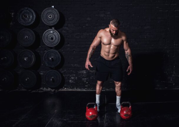 A man stands confidently, holding a kettlebell in each hand, ready to engage in a powerful workout session. - Fitsse